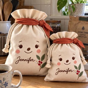 May include: Two off-white drawstring pouches made of fabric, each featuring an embroidered smiling face with pink cheeks, the name Jennifer in cursive script, and a small botanical sprig with red berries and a white flower, tied with a burnt orange ribbon.