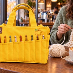 Op de afbeelding: Een felgele canvas knutseltas met twee handvatten en een reeks buitenvakken met diverse gekleurde haaknaalden. De tas is voorzien van een geborduurd oranje pompoenontwerp en een klein bruin leren logolabel met de letters CC.