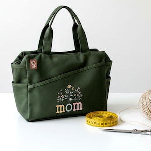 Op de afbeelding: Een donker olijfgroene canvas tas met twee handvatten, zijvakken en een klein bruin rechthoekig merklabel. De voorzijde toont een bloemmotief boven de tekst "mom" in pastelkleurige letters, naast een geel meetlint op de ondergrond.