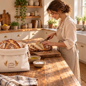 May include: A cream-coloured fabric bread bag with a drawstring closure, featuring an embroidered design of a loaf of bread flanked by two wheat stalks and the name Emma printed in brown text on the front, holding a round loaf of crusty bread.