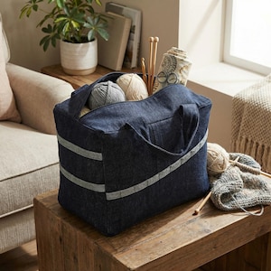 Op de afbeelding: Een donkerblauwe denim opbergtas met twee horizontale lichtgrijze stoffen banden aan de voorzijde. De tas is gevuld met diverse bollen garen in grijze en crèmetinten, samen met houten breinaalden en een rol met bedrukte stof.