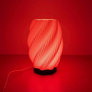 Pode incluir: Uma lâmpada de mesa vermelha com um padrão em espiral e uma base preta. A lâmpada está iluminada, projetando um brilho vermelho quente. O design da lâmpada apresenta linhas verticais que criam um efeito texturizado. Uma luz vermelha brilha no teto.