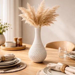Può includere: Un vaso di ceramica bianca con un motivo strutturato contiene un bouquet di erba della pampa essiccata. Il vaso è su un tavolo di legno, insieme a piatti, tovaglioli e un bicchiere d'acqua. La scena suggerisce un tema di sala da pranzo o arredamento.