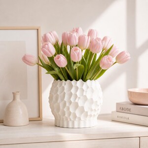 May include: A white textured vase filled with pink tulips. The vase has a unique geometric pattern and sits on a white surface. A small, light-colored vase and framed artwork are in the background.