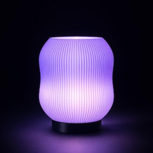 以下が含まれることがあります： 紫色のモダンなランプ。リブ加工が施された丸みを帯びたデザインです。柔らかく拡散した光を放ち、落ち着いた雰囲気を演出します。ベースは黒で、光る本体とコントラストをなしています。インテリアに最適です。