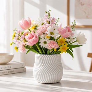 Puede incluir: Un vibrante ramo de flores primaverales en un jarrón blanco texturizado. El arreglo incluye tulipanes rosas, margaritas blancas, narcisos amarillos y otras flores coloridas. El jarrón tiene un diseño ondulado.