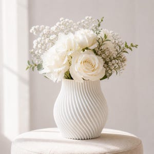 May include: A white ceramic vase with a spiral design holds a bouquet of white roses, peonies, and baby's breath. The arrangement is set on a neutral-toned surface, creating a soft, elegant aesthetic. The vase is approximately 8 inches tall.