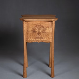 Puede incluir: Una mesita de noche de madera marrón claro con un grabado de árbol en el frente del cajón. La mesita tiene una parte superior rectangular con bordes curvos, dos cajones y cuatro patas cónicas. El grabado del árbol es detallado y centrado.