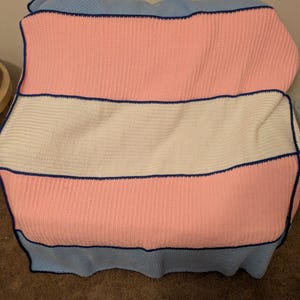 Könnte beinhalten: Eine handgefertigte Decke in den Farben der Transgender-Pride-Flagge. Die Decke hat horizontale Streifen in Rosa, Weiß und Hellblau mit einem dunkelblauen Rand. Die Decke besteht aus einem strukturierten Strickmaterial.