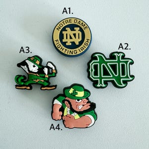 May include: Four Notre Dame Fighting Irish themed shoe charms. A1 is a circular gold and navy emblem. A2 is a green and black interlocking ND logo. A3 is a green and gold leprechaun figure in a fighting stance. A4 is a green and tan leprechaun head.