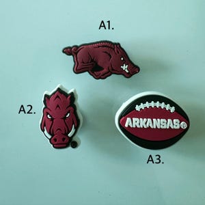 May include: Three shoe charms labeled A1, A2, and A3. A1 is a maroon running razorback hog. A2 is a maroon razorback hog head. A3 is an oval-shaped football design in maroon, black, and white with the text ARKANSAS and a registered trademark symbol.