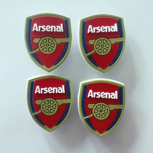 May include: Four identical shield-shaped charms featuring the Arsenal football club logo. Each emblem displays a red background with a gold cannon graphic, blue trim, and the word Arsenal written in white capital letters across the top section.
