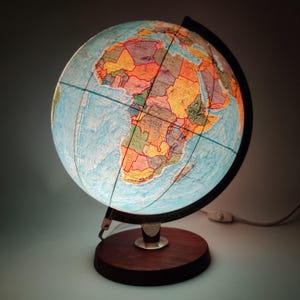 Op de afbeelding: Verlichte wereldbol met een gedetailleerde wereldkaart, met continenten en landen in verschillende kleuren. De bol staat op een donkere houten voet met een metalen steun. Een netsnoer met een aan/uit-schakelaar is bevestigd.