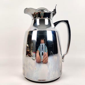 Könnte beinhalten: Eine glänzende, verchromte Getränkekaraffe mit gebogenem Griff und Klappdeckel. Die Karaffe hat einen Ausguss zum Ausgießen und einen schwarzen Sockel. Die Oberfläche reflektiert die Umgebung.