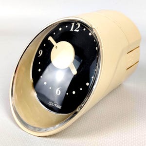 Op de afbeelding: Een crèmekleurige vintage wekker met een zwarte wijzerplaat en witte stipuurmarkeringen. De wijzerplaat heeft de cijfers 12, 9 en 6 in het wit. De klok heeft een gebogen, ovale vorm met een zilverkleurige rand en de tekst "EDUTIME" is zichtbaar.