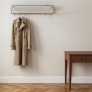 Puede incluir: Un abrigo tipo trench de color beige cuelga de un perchero de pared. El abrigo tiene un diseño cruzado y un cinturón. Una mesa auxiliar de madera con un cajón está a la derecha de la imagen. El suelo tiene un patrón de espiga.