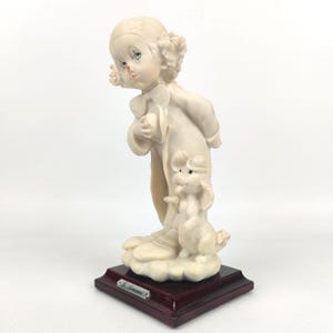 Könnte beinhalten: Porzellanfigur eines Clowns mit einem kleinen Hund. Der Clown trägt ein weißes Outfit und einen passenden Hut mit bemalten Gesichtszügen. Der Hund steht neben dem Clown. Die Figur ist auf einem dunklen Holzsockel montiert.