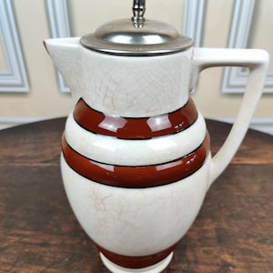 May include: A vintage ceramic jug with a white and brown striped design. The jug has a silver lid and handle. The jug is approximately 18 cm tall.