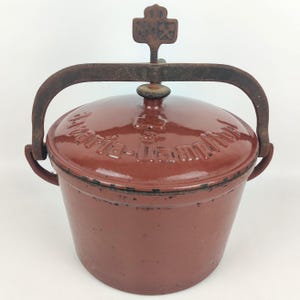 Op de afbeelding: Een rode gietijzeren pot met een deksel en een handvat. Op het deksel staan de woorden "Marie-Jeanne" in reliëf. De pot heeft een handvat en een schroefmechanisme aan de bovenkant. De pot wordt waarschijnlijk gebruikt om te koken.