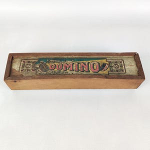 Könnte beinhalten: Eine rechteckige Dominobox aus Holz mit dem Wort "DOMINO" auf dem Deckel. Die hellbraune Box hat ein dekoratives Design. Sie dient wahrscheinlich zur Aufbewahrung von Dominosteinen, einem klassischen Spiel.