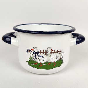 Op de afbeelding: Witte emaille pot met een donkerblauwe rand en handvatten. De pot heeft een charmante illustratie van twee eenden met blauwe strikken, lopend bij een hek en groen gras. Een decoratief keukenartikel.