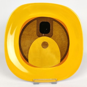 Könnte beinhalten: Ein quadratischer, gelber Zierteller mit einem kreisförmigen Design in Braun- und Gelbtönen. Das Design umfasst ein schwarzes Quadrat und einen gelben Kreis. Der Teller wird auf einem transparenten Ständer präsentiert.