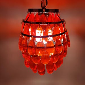 Puede incluir: Una lámpara de araña decorativa con una estructura cromada y cristales en forma de lágrima de color naranja-rojo. La lámpara está suspendida de una cadena y emite un brillo cálido, creando una exhibición vibrante y llamativa.