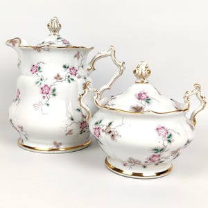 Può includere: Un servizio di panna e zuccheriera in porcellana bianca. Entrambi i pezzi presentano un motivo floreale con fiori rosa e foglie verdi, rifinito con bordi dorati. La panna ha un manico e un beccuccio curvi.