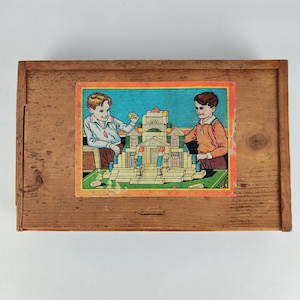 May include: A vintage wooden toy box featuring a colourful illustration of two children constructing a building with blocks. The rectangular box is crafted from light brown wood, highlighting the age and character of the toy.