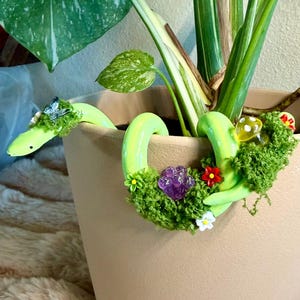 May include: A tan ceramic plant pot with a Monstera plant. A light green, whimsical snake-shaped plant pot decoration is wrapped around the rim, adorned with moss, flowers, and a purple crystal. The snake's head is near the plant's leaves.