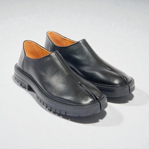 Könnte beinhalten: Ein Paar schwarze Lederschlupfschuhe mit geteiltem Zehendesign. Die Schuhe haben eine dicke Profilsohle und ein glattes Obermaterial. Der Innenraum ist mit hellbraunem Leder gefüttert.