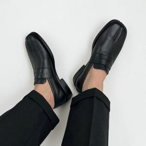 May include: A pair of black leather loafers with a square toe design. The shoes have a low heel and a strap detail across the top. The image shows the shoes from a top-down perspective, with the wearer's black trousers visible.