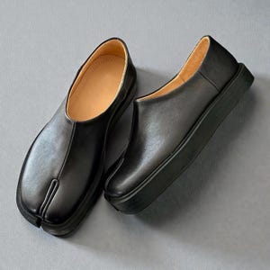 Könnte beinhalten: Ein Paar schwarze Lederschlupfschuhe mit geteiltem Zehendesign. Die Schuhe haben eine dicke, schwarze Sohle und ein beiges Innenfutter. Die Schuhe werden auf grauem Hintergrund präsentiert.
