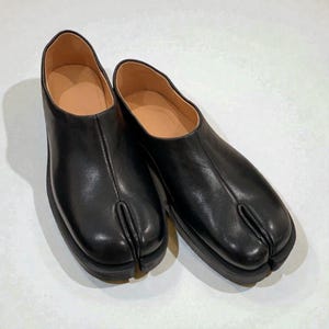 Könnte beinhalten: Ein Paar schwarze Lederschuhe mit geteilter Zehenpartie und dicker Sohle. Die Schuhe haben ein einzigartiges Design mit einem separaten Fach für den großen Zeh. Die Innenseite ist hellbraun.