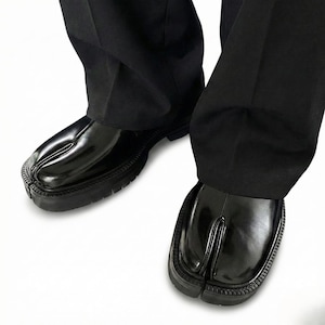 May include: Black split-toe shoes with a shiny surface and chunky, ridged soles. The shoes are worn with black trousers, forming a monochrome outfit. The design includes a distinctive split between the big toe and the other toes.