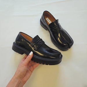 Op de afbeelding: Zwarte leren split-toe loafers met een dikke zool. De schoenen hebben een glanzende afwerking en een slip-on ontwerp met een band over de bovenkant. De zool is ongeveer 3,8 cm dik. De binnenkant van de schoen heeft een label met "ArukuTabi".