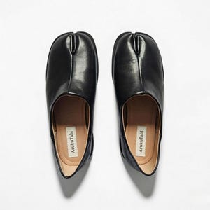 Könnte beinhalten: Eine weiße Keramik-Tasse mit schwarzem Henkel und dem Text "But First, Coffee" in fetter, schwarzer Schrift. Die Tasse steht vor einem verschwommenen Hintergrund, der eine gemütliche Umgebung suggeriert.