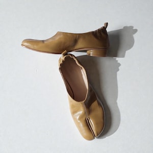 Puede incluir: Dos pares de zapatos de cuero color canela sobre un fondo blanco. Uno es un zapato plano con tacón bajo. El otro es una bailarina con punta dividida. Los zapatos están en ángulo, creando sombras.