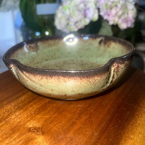 May include: A shallow ceramic bowl with a light green interior and exterior featuring brown speckled details. The rim is dark brown and includes two small indentations on opposite sides, creating a distinct shape with a textured, earthy finish.