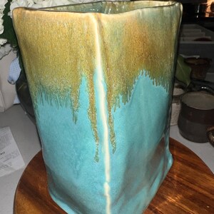 May include: A rectangular ceramic vase with a textured, uneven surface. The upper portion features a mottled golden-brown glaze that drips down into a vibrant turquoise blue glaze covering the lower section, creating a distinct color transition.