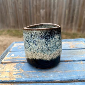 Puede incluir: Una taza de cerámica hecha a mano con un esmalte texturizado único. La taza presenta una base azul oscuro que se desvanece en un patrón moteado azul claro y beige, con un borde marrón oscuro. La taza tiene una forma ligeramente irregular.