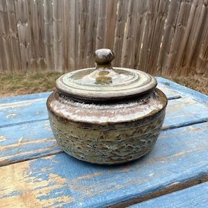 May include: A handcrafted ceramic jar with a lid. The jar is a mix of earthy tones, including brown, green, and beige, with a textured surface. The lid has a small knob on top. The jar sits on a weathered blue surface.
