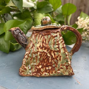 May include: A handmade ceramic teapot with a textured surface. The teapot features a triangular shape with a brown, green, and tan color scheme. The handle and spout have a rustic, bark-like appearance, and the lid is topped with a small knob.