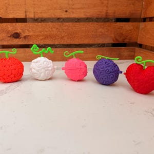 May include: Five colorful, stylized fruit-shaped objects with green stems. The fruits are red, white, pink, purple, and red heart-shaped. Each has a unique textured surface and a small, red protrusion. The objects are arranged on a white surface.