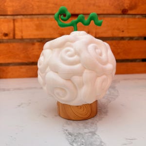 May include: A white, cloud-shaped object with a green, swirly stem, resting on a wooden base. The cloud has a textured, swirling design. The base is a light brown color. The background is a wooden wall.