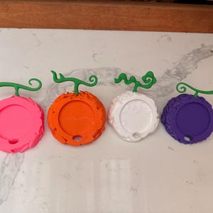 May include: Four colorful, round, 3D-printed objects resembling stylized fruits. The fruits are in pink, orange, white, and purple, each with a green stem. The objects have a circular design with a hole in the bottom.
