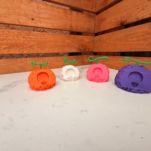 May include: Four colorful, stylized fruit-shaped objects with green stems. The fruits are orange, white, pink, and purple, each with a circular opening. They are arranged on a white surface against a wooden background.