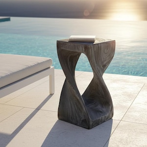 May include: A gray, sculptural side table with a twisted design, topped with a white book. The table is made of a textured material and is placed near a pool and lounge chair, with the ocean in the background.