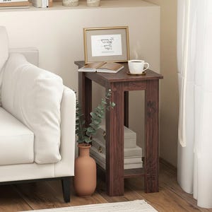 May include: A dark brown wooden side table with a rectangular top and open sides. A framed print, a cup of coffee, and a book rest on top. A vase with greenery and stacked books are on the lower shelf. A white sofa is on the left.