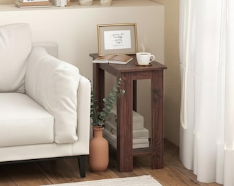 May include: A dark brown wooden side table with a rectangular top and open sides. A framed print, a cup of coffee, and a book rest on top. A vase with greenery and stacked books are on the lower shelf. A white sofa is on the left.
